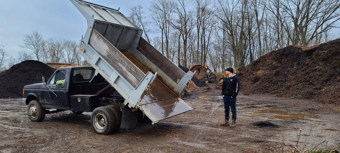 Man standing near a dump truck unloading soil at a muddy compost or mulch yard with large soil piles