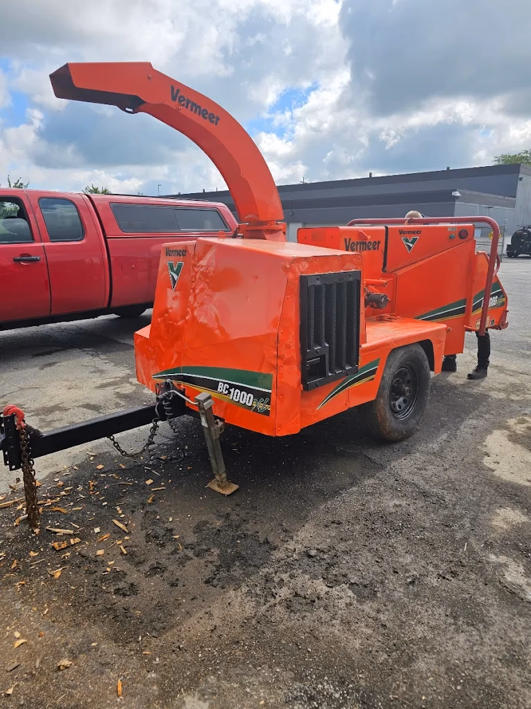 Orange Vermeer BC1000 wood chipper machine parked on asphalt near a red pickup truck.