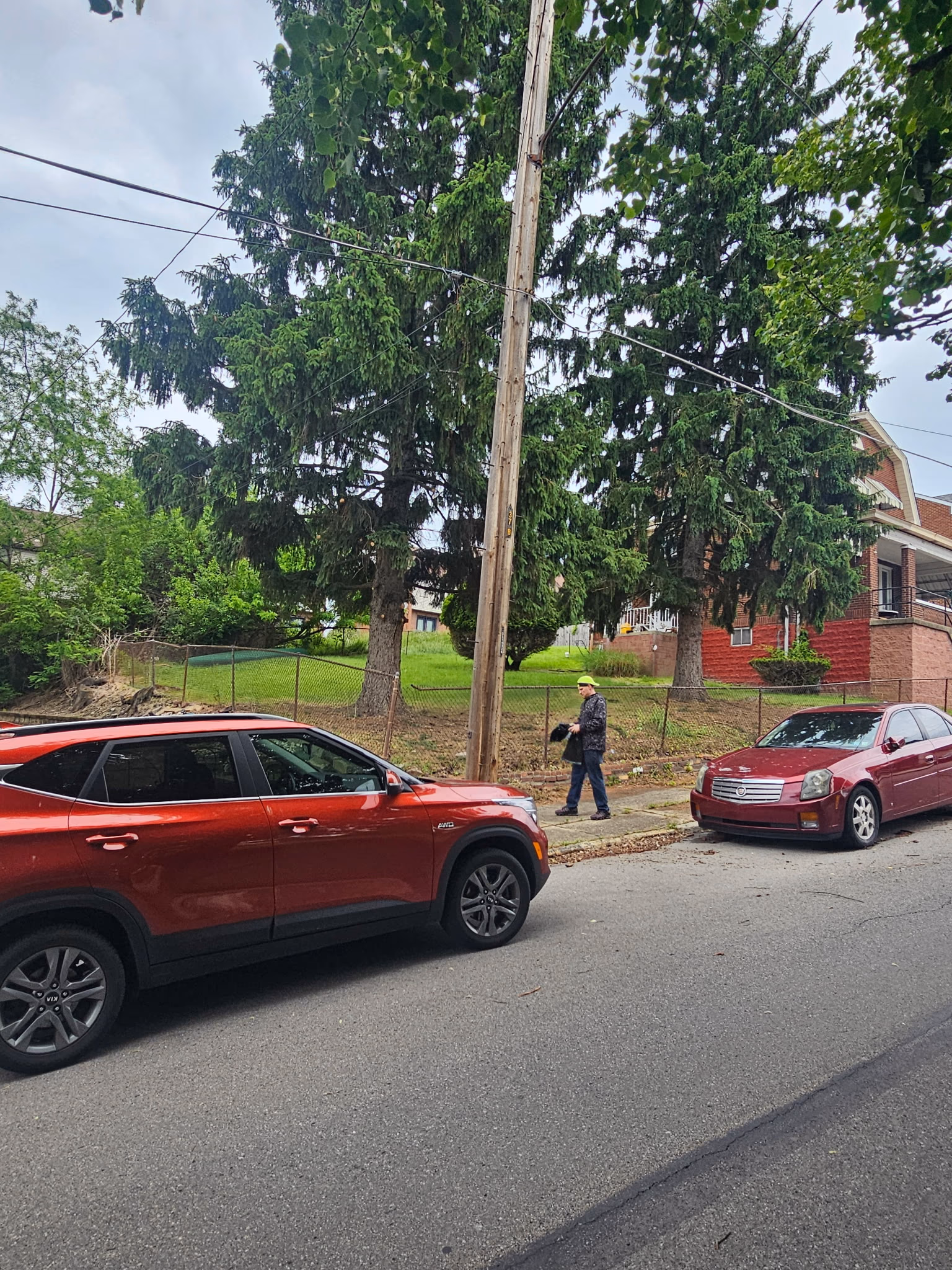 Man in a yellow cap walking on a sidewalk near two red cars parked on the street with trees behind.