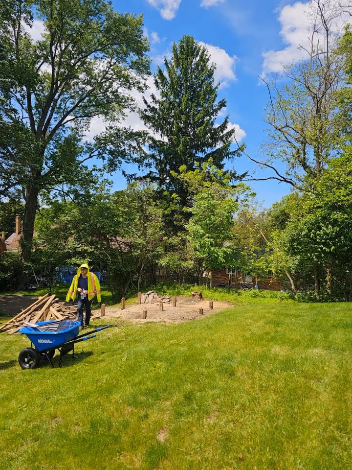 Worker in yellow vest and hoodie standing near blue wheelbarrow and wooden posts in grassy backyard.