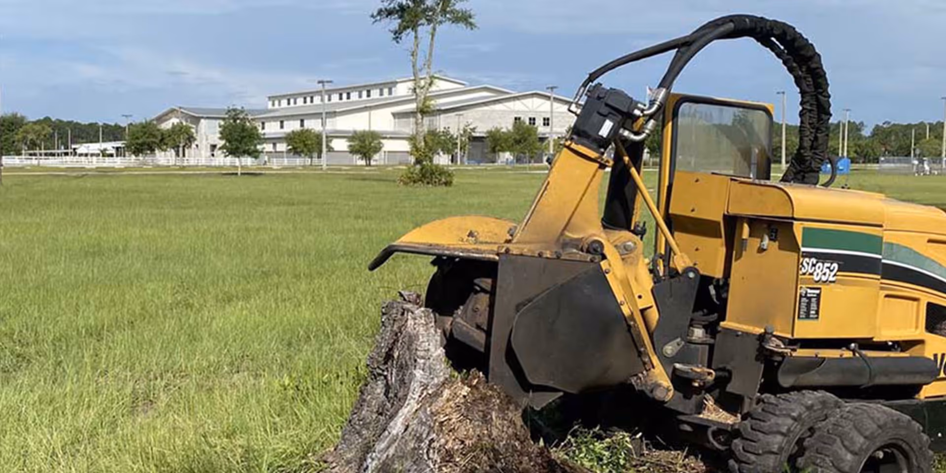 Yellow stump grinder machine attached to a large tree stump in a grassy field with buildings in background.